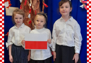 Maja,Paulinka,Magda w odświętnym ubraniu trzymają w rączkach flagę Polski, w tle dekoracja okolicznościowa